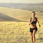 Powerful young female athlete in activewear running along hill on background of mountainous landscape and listening to music in earphones during cardio training